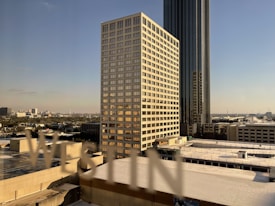 A cityscape featuring a tall, modern skyscraper with a glass facade and a smaller, rectangular building with numerous windows. The sky is clear, suggesting a likely sunny day. The letters 'WESTIN' appear in the foreground, possibly seen through a window.