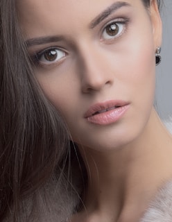 A close-up portrait of a person with smooth, flawless skin and long brown hair. The focus is on the face, highlighting large, expressive brown eyes and slightly parted lips with a natural look. The person is wearing subtle eye makeup and an earring is visible in one ear.