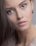 A close-up portrait of a person with smooth, flawless skin and long brown hair. The focus is on the face, highlighting large, expressive brown eyes and slightly parted lips with a natural look. The person is wearing subtle eye makeup and an earring is visible in one ear.