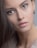 A close-up portrait of a person with smooth, flawless skin and long brown hair. The focus is on the face, highlighting large, expressive brown eyes and slightly parted lips with a natural look. The person is wearing subtle eye makeup and an earring is visible in one ear.