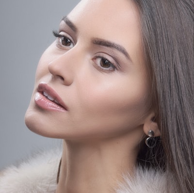 A close-up portrait of a woman with smooth, flawless skin and neutral makeup, featuring well-defined eyebrows and slightly glossy lips. Her hair is straight and drapes smoothly along the side of her face. She is wearing a pair of simple black stud earrings and a fur collar, with a soft, neutral grey background.