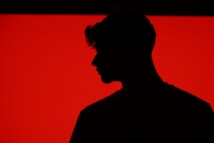 A silhouette of a person with a distinct hairstyle is set against a bold, bright red background. The figure is in profile, with details obscured by shadow, creating a stark contrast with the vivid backdrop.
