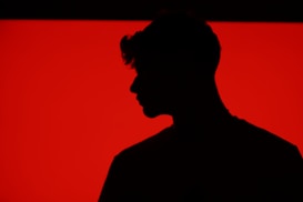 A silhouette of a person with a distinct hairstyle is set against a bold, bright red background. The figure is in profile, with details obscured by shadow, creating a stark contrast with the vivid backdrop.
