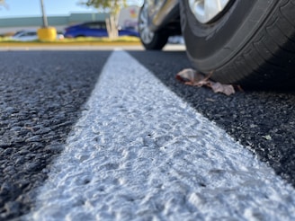 Close-up of a car tire on a textured asphalt surface with a prominent white line running through the center. The view is from ground level, and there are autumn leaves near the tire. In the background, there are blurred images of other parked cars, trees, and a large yellow object.