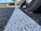 Close-up of a car tire on a textured asphalt surface with a prominent white line running through the center. The view is from ground level, and there are autumn leaves near the tire. In the background, there are blurred images of other parked cars, trees, and a large yellow object.