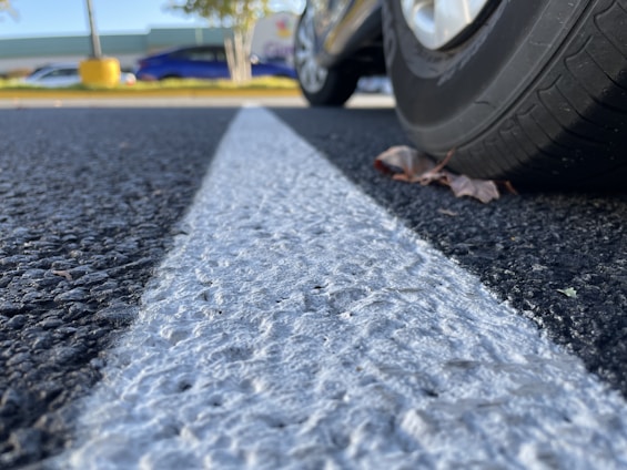 Close-up of a car tire on a textured asphalt surface with a prominent white line running through the center. The view is from ground level, and there are autumn leaves near the tire. In the background, there are blurred images of other parked cars, trees, and a large yellow object.