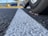 Close-up of a car tire on a textured asphalt surface with a prominent white line running through the center. The view is from ground level, and there are autumn leaves near the tire. In the background, there are blurred images of other parked cars, trees, and a large yellow object.