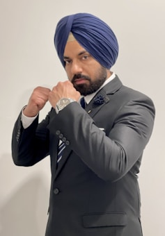 A man in a formal suit poses with a confident stance, wearing a blue turban. His hands are raised in a boxing guard position. The suit includes a tie and a flower lapel pin, and he has a watch on his left wrist.