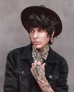 A person with dark hair wearing a black hat, nose rings, and earrings. The individual has several tattoos, including one with a detailed design on the hand. A plaid shirt is visible under a dark denim jacket, and the person is looking to the side.