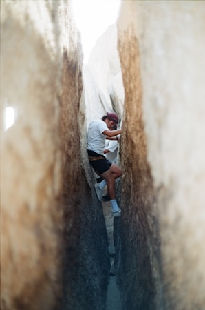 A person climbs between two large boulders, using their arms and legs to brace against the walls. They are wearing a white shirt, dark shorts, socks, and sneakers, with a cap on their head. The rocks have a rough texture and range in color from tan to gray, with sunlight filtering through the narrow passage.