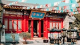 A traditional Chinese temple with vibrant red and blue detailing. The building features intricate window designs and decorative elements. Colorful prayer flags are strung across the scene, adding a dynamic aspect to the image. In front of the temple are two ornate black incense burners.
