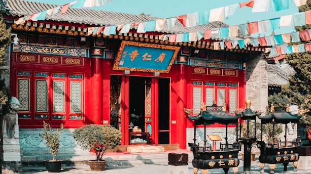 A traditional Chinese temple with vibrant red and blue detailing. The building features intricate window designs and decorative elements. Colorful prayer flags are strung across the scene, adding a dynamic aspect to the image. In front of the temple are two ornate black incense burners.
