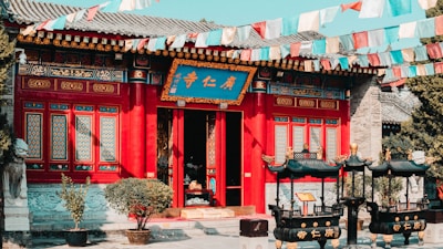 A traditional Chinese temple with vibrant red and blue detailing. The building features intricate window designs and decorative elements. Colorful prayer flags are strung across the scene, adding a dynamic aspect to the image. In front of the temple are two ornate black incense burners.
