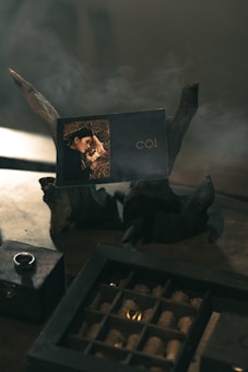 A card with the name 'cội' placed on a piece of dark driftwood surrounded by smoke or mist. Nearby, there is a small, closed wooden box with a ring on top and a larger compartmentalized box containing multiple pine cones.