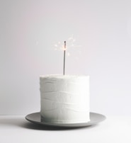 A minimalist white cake with a smooth frosting texture sits on a gray plate. A lit sparkler is placed on top of the cake, emitting small sparks.