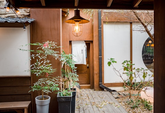 A serene courtyard with wooden elements and minimalistic decor. There are two potted plants with green foliage and red berries. A door with a white banner hangs in the center. The courtyard is enclosed with walls featuring both brown and white panels. A soft light fixture is hanging above, casting a warm glow.