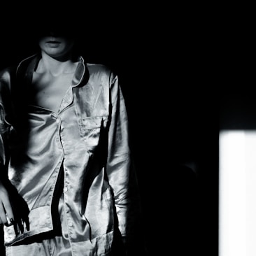 A person wearing a satin shirt stands partially in shadow with their face obscured. The lighting creates a dramatic contrast between the illuminated fabric and the dark background.