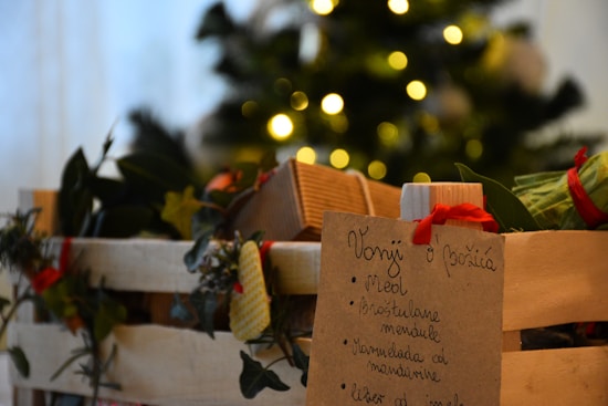 A wooden basket filled with festive items, including greenery and wrapped gifts. There's a handwritten list on a piece of paper attached to the basket, containing items possibly associated with Christmas. In the blurred background, a decorated Christmas tree with glowing lights adds to the holiday ambiance.