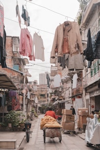 A narrow alleyway lined with residential buildings features clothes hanging overhead on lines. A person in a red jacket is pushing a cart loaded with goods, and another person stands nearby, surrounded by cardboard boxes and parcels. The scene suggests a bustling urban environment with activity and commerce, emphasized by the laundry adding a lively touch to the setting.