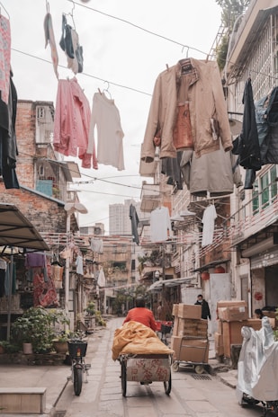 A narrow alleyway lined with residential buildings features clothes hanging overhead on lines. A person in a red jacket is pushing a cart loaded with goods, and another person stands nearby, surrounded by cardboard boxes and parcels. The scene suggests a bustling urban environment with activity and commerce, emphasized by the laundry adding a lively touch to the setting.