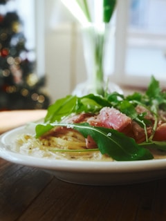 A plate of pasta garnished with arugula leaves and slices of pink-cooked meat sits on a wooden table. The background features a softly lit room with a blurred Christmas tree adorned with lights, creating a cozy and festive ambiance.