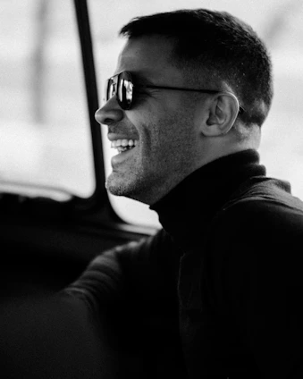 Portrait of Jorge Villegas smiling warmly in front of a sleek luxury car with a subtle black and deep red gradient background.