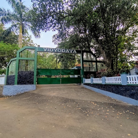 A large green gate with 'VIDYODAYA' written overhead is flanked by green metal fencing and tall trees. A pathway leads up to the entrance, surrounded by white fencing and lush greenery.