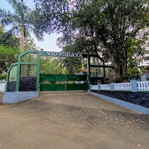 A large green gate with 'VIDYODAYA' written overhead is flanked by green metal fencing and tall trees. A pathway leads up to the entrance, surrounded by white fencing and lush greenery.