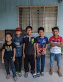 Five boys stand in a row against a blue wall with a window behind them. Each is wearing casual clothing, including t-shirts and sandals. They appear to be smiling and relaxed.
