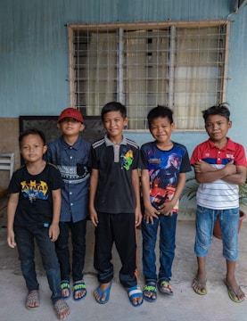 Five boys stand in a row against a blue wall with a window behind them. Each is wearing casual clothing, including t-shirts and sandals. They appear to be smiling and relaxed.