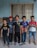 Five boys stand in a row against a blue wall with a window behind them. Each is wearing casual clothing, including t-shirts and sandals. They appear to be smiling and relaxed.