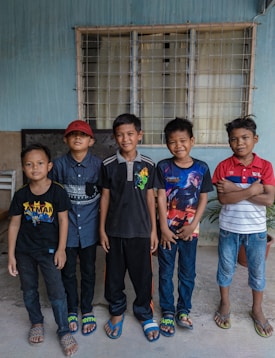 Five boys stand in a row against a blue wall with a window behind them. Each is wearing casual clothing, including t-shirts and sandals. They appear to be smiling and relaxed.