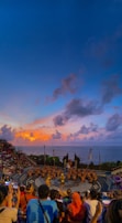 A large audience watches a traditional dance performance taking place in an amphitheater overlooking the ocean. The performers are in the center, wearing costumes, while the sky above is filled with vibrant colors from the setting sun.