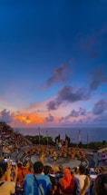 A large audience watches a traditional dance performance taking place in an amphitheater overlooking the ocean. The performers are in the center, wearing costumes, while the sky above is filled with vibrant colors from the setting sun.