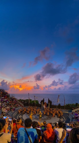A large audience watches a traditional dance performance taking place in an amphitheater overlooking the ocean. The performers are in the center, wearing costumes, while the sky above is filled with vibrant colors from the setting sun.