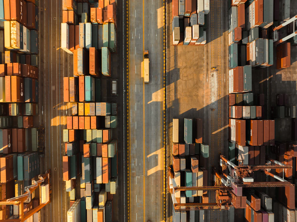 The image presents an aerial view of a bustling port, teeming with activity and filled with stacks of shipping containers. The containers vary in size and color, creating a vibrant mosaic against the backdrop of the port's infrastructure. A road runs through the center of the image, serving as a vital artery for the movement of goods. On this road, a yellow truck is seen driving past the towering stacks of containers.
