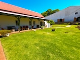 A well-maintained garden features a lush, green lawn adjacent to a building with a red corrugated iron roof and white walls. The building has a porch area furnished with chairs and tables. In the background, another larger building with a white facade and arched doorways is visible. The garden includes various potted plants and a few scattered decorative items.