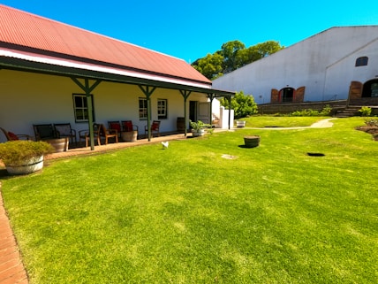 A well-maintained garden features a lush, green lawn adjacent to a building with a red corrugated iron roof and white walls. The building has a porch area furnished with chairs and tables. In the background, another larger building with a white facade and arched doorways is visible. The garden includes various potted plants and a few scattered decorative items.