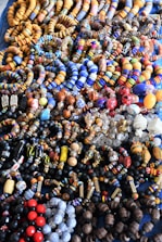 An assortment of colorful beaded jewelry, including bracelets and necklaces, laid out in a densely packed display. The beads vary in shape, size, and pattern, featuring a vibrant array of colors such as blue, orange, red, and black. The intricate designs and variety suggest hand-crafted or artisanal origins.
