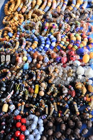An assortment of colorful beaded jewelry, including bracelets and necklaces, laid out in a densely packed display. The beads vary in shape, size, and pattern, featuring a vibrant array of colors such as blue, orange, red, and black. The intricate designs and variety suggest hand-crafted or artisanal origins.