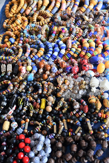 An assortment of colorful beaded jewelry, including bracelets and necklaces, laid out in a densely packed display. The beads vary in shape, size, and pattern, featuring a vibrant array of colors such as blue, orange, red, and black. The intricate designs and variety suggest hand-crafted or artisanal origins.