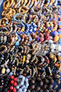 An assortment of colorful beaded jewelry, including bracelets and necklaces, laid out in a densely packed display. The beads vary in shape, size, and pattern, featuring a vibrant array of colors such as blue, orange, red, and black. The intricate designs and variety suggest hand-crafted or artisanal origins.
