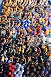 An assortment of colorful beaded jewelry, including bracelets and necklaces, laid out in a densely packed display. The beads vary in shape, size, and pattern, featuring a vibrant array of colors such as blue, orange, red, and black. The intricate designs and variety suggest hand-crafted or artisanal origins.