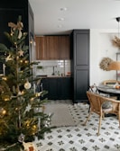 A cozy kitchen with dark wood cabinets and black countertops against a white subway tile backsplash. A decorated Christmas tree with lights and ornaments is positioned in the foreground. The floor features geometric patterned tiles, and a dining area with rattan chairs and a round table is visible. Warm lighting and natural elements like hanging dried plants create a welcoming atmosphere.