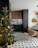 A cozy kitchen with dark wood cabinets and black countertops against a white subway tile backsplash. A decorated Christmas tree with lights and ornaments is positioned in the foreground. The floor features geometric patterned tiles, and a dining area with rattan chairs and a round table is visible. Warm lighting and natural elements like hanging dried plants create a welcoming atmosphere.
