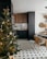 A cozy kitchen with dark wood cabinets and black countertops against a white subway tile backsplash. A decorated Christmas tree with lights and ornaments is positioned in the foreground. The floor features geometric patterned tiles, and a dining area with rattan chairs and a round table is visible. Warm lighting and natural elements like hanging dried plants create a welcoming atmosphere.