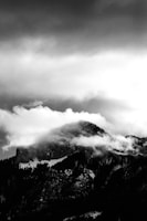 A dramatic black and white photograph of a mountain shrouded in clouds. The rugged mountain peak is partially covered by swirling mist, creating an ethereal and mysterious atmosphere. The dark silhouette of the mountain contrasts with the bright, diffused light from the clouds above.