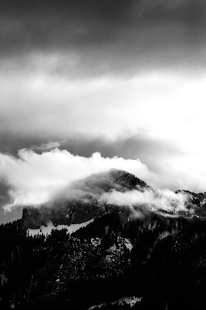 A dramatic black and white photograph of a mountain shrouded in clouds. The rugged mountain peak is partially covered by swirling mist, creating an ethereal and mysterious atmosphere. The dark silhouette of the mountain contrasts with the bright, diffused light from the clouds above.