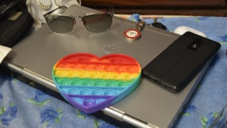 A silver laptop is placed on a blue cloth with floral patterns. Resting on the laptop is a pair of sunglasses and a round emblem keychain. A black smartphone lies beside the laptop. In front of the laptop, a colorful heart-shaped pop-it toy with rainbow stripes is prominently visible.