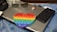 A silver laptop is placed on a blue cloth with floral patterns. Resting on the laptop is a pair of sunglasses and a round emblem keychain. A black smartphone lies beside the laptop. In front of the laptop, a colorful heart-shaped pop-it toy with rainbow stripes is prominently visible.